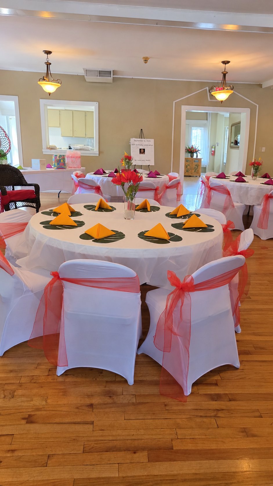 Event space with rectangular tables draped in white linens and white chair covers tied with pink organza sashes, dressed with floral centerpieces